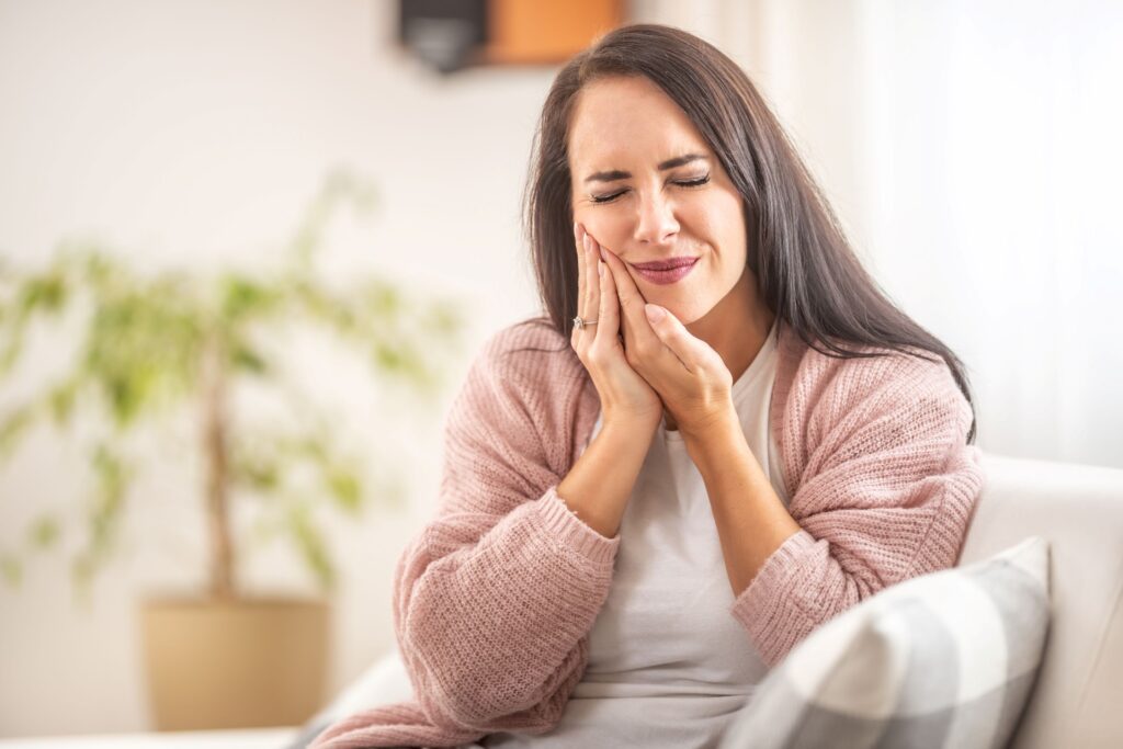 Woman sitting on couch in pink sweater holding hands to jaw in pain